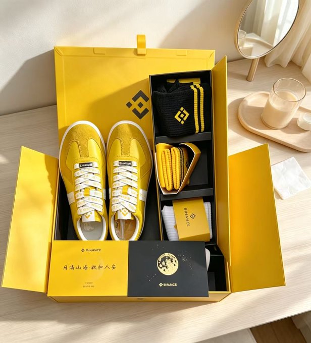 An open yellow Binance promotional box displays branded yellow and white sneakers, a black beanie with the Binance logo, yellow shoelaces, socks, and an envelope.