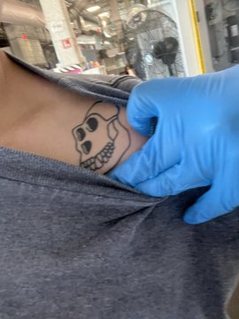 A gloved hand stretches the fabric of a gray shirt to reveal a small tattoo of a cartoon skull character on someone's skin.