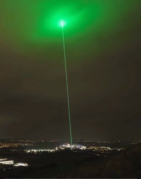 A nighttime photograph showing a bright green laser beam shooting into the sky from a city below, with a glowing green cloud at its apex.