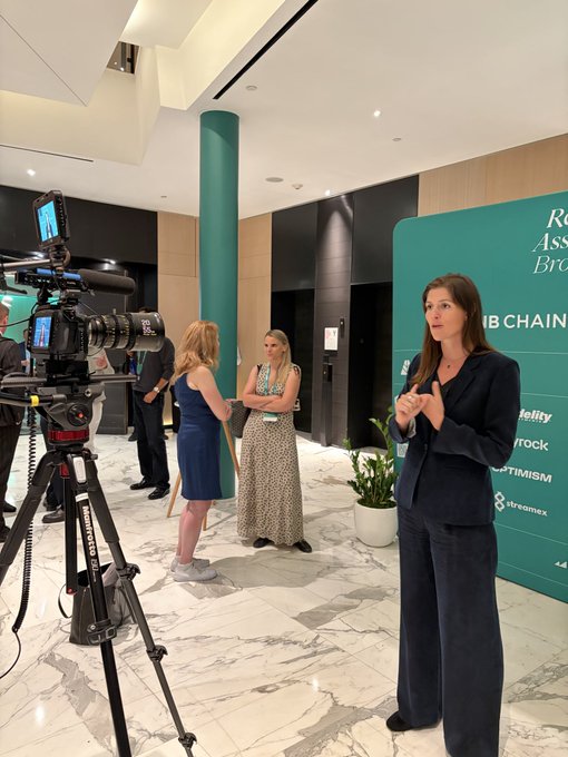 A woman in a dark blazer gives an interview in a lobby while a camera on a Sachtler tripod films her; a backdrop behind her reads "ROBINHOOD CHAIN" with text fragments like "stellity," "ntock," and "PTIMISM."