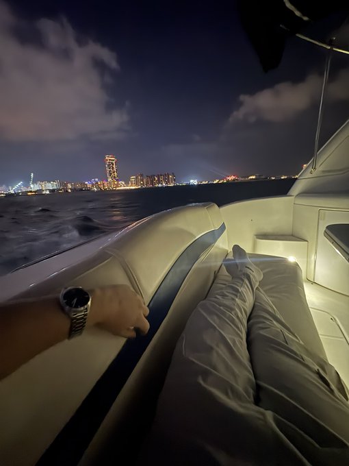 A nighttime photo taken from a boat showing a person's legs resting on a cushioned seat, their wrist displaying a luxury watch, with a glowing city skyline in the distance across the water.