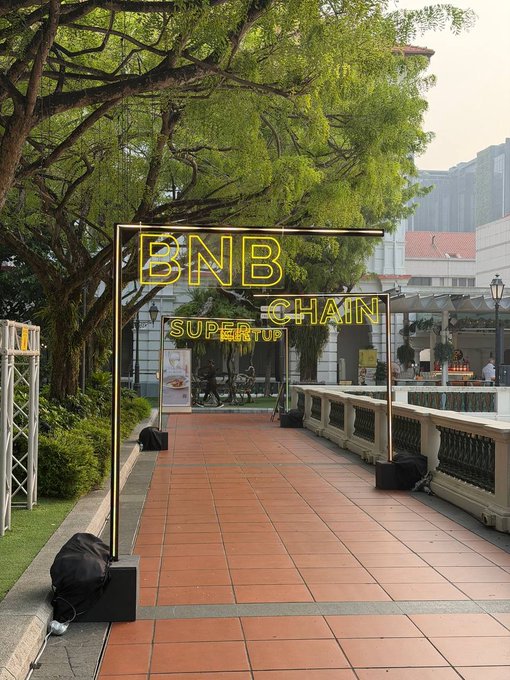 An outdoor event setup featuring a neon sign reading 'BNB SUPER CHAIN STARTUP' over a tiled walkway, with a building and food stall in the distance.
