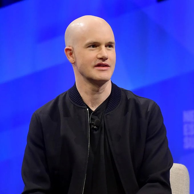 A bald man in a black bomber jacket speaks at what appears to be a conference, likely Coinbase CEO Brian Armstrong based on the accompanying context about his comments on stablecoins and their adoption in high-inflation countries.