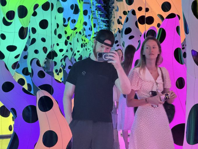 A man in a black shirt and backwards cap takes a mirror selfie with his phone while a woman in a pink polka-dot dress stands beside him in a colorful installation featuring large polka-dotted shapes in vibrant colors.