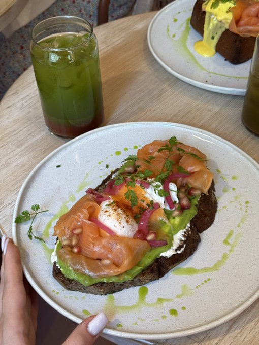 A close-up of a café meal featuring a slice of dark bread topped with avocado, smoked salmon, pickled onions, pine nuts, and a poached egg, served alongside a green iced drink.