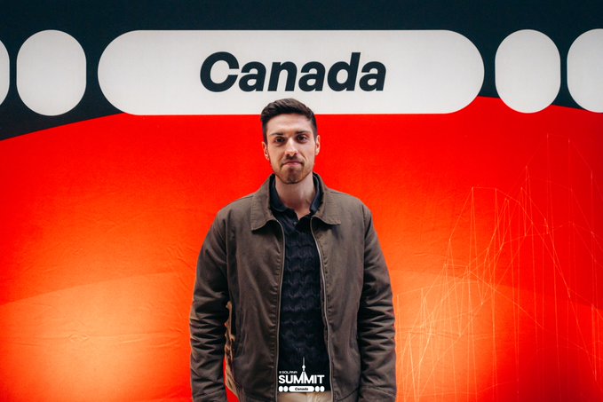 A man in a brown jacket poses in front of the same red and white 'Canada' and 'Summit Toronto' backdrop.