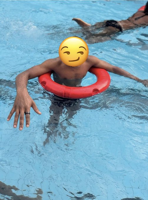A person is in a swimming pool using a red floating ring, with their face covered by a smiley emoji sticker.