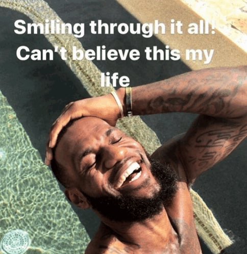 The image features a man with tattoos, wearing no shirt or bathing suit, smiling while leaning over a pool of water. The caption below the photo reads "Smiling through it all! Can't believe this my life", which seems to be related to the content of the image.
