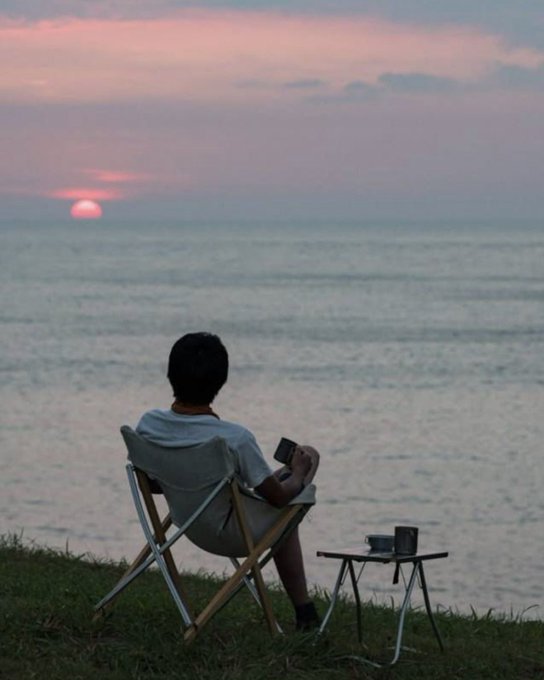 A silhouetted person sitting in a folding chair on grass at the seaside, watching the sunset while holding a cup, with a small table beside them.