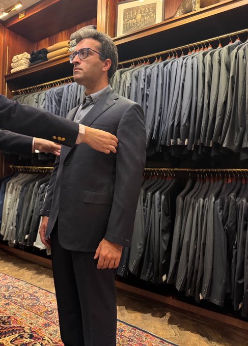 A man being fitted for a dark suit jacket in a tailor shop with rows of hanging suits in the background.