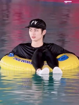 A young man wearing a black cap with "PV" text and a black long-sleeved shirt sits in a yellow "SWIM RING" inflatable tube in a pool. The image appears to promote the hashtagged artists and the ALPHADRIVEONE debut, possibly featuring Zhou Anxin or a related member of the K-pop group.
