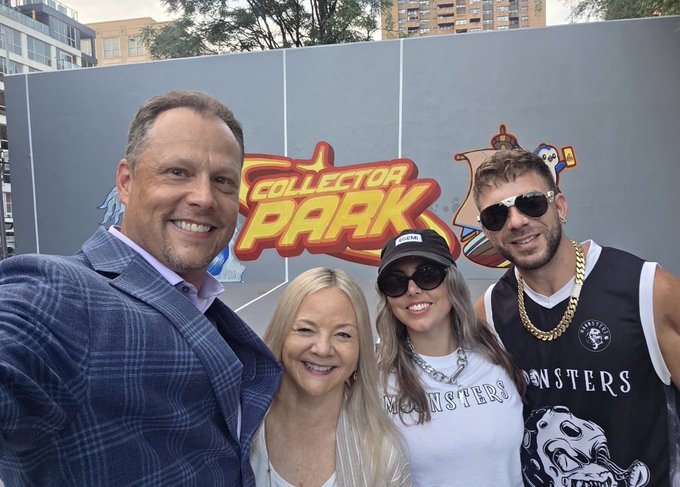A selfie of four people smiling in front of a 'COLLECTOR PARK' sign, with two wearing matching 'MONSTERS' shirts and accessories.