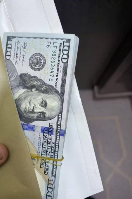 A stack of $100 U.S. bills bound with a rubber band, featuring the serial number "LF 38263647 E," is being pulled from a manila envelope—a visual nod to the hustle and grind of chasing money.