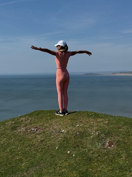 A woman wearing a pink athletic outfit and a white cap stands with her arms outstretched on a grassy cliff overlooking the ocean under a clear blue sky.