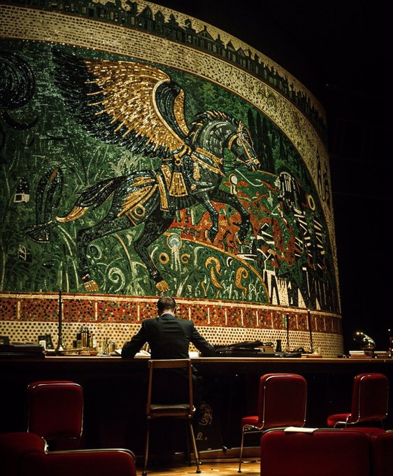 A man in a dark suit sits alone at a table in an ornate, dimly-lit hall, facing a massive, vividly colored mosaic mural depicting a winged horse.