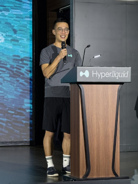 A young man in a gray t-shirt and shorts stands at a podium labeled "Hyperliquid," holding a microphone as he addresses an audience. The setting aligns with the context of Hyperliquid co-founder Jeff Yan speaking at Token2049 Singapore, presenting the platform as an open financial tool rather than a competitor.