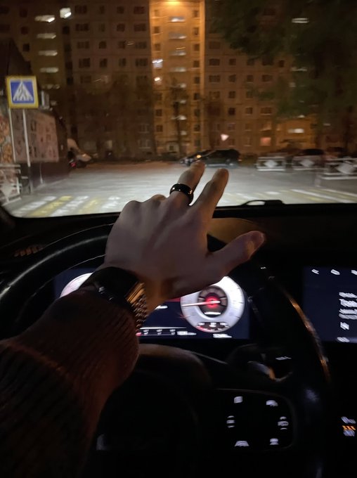 A first-person view from inside a car at night, with a hand wearing a watch and ring resting on the steering wheel, dashboard displays illuminated, and apartment buildings visible ahead through the windshield.