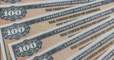 The image displays a stack of United States Treasury 100 bills with the text "United States Savings Bond" printed on them. The bills appear to be new and fresh, suggesting that they have not been used or minted recently. They are organized in several piles, one at each corner, all facing upwards against a brown background.
