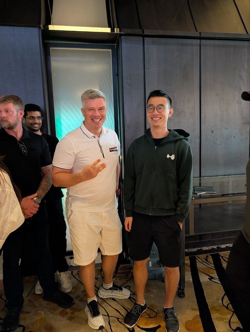 Two men pose smiling at an indoor event, one in a white polo shirt holding a chip and the other in a green hoodie with glasses, with other attendees visible in the background.