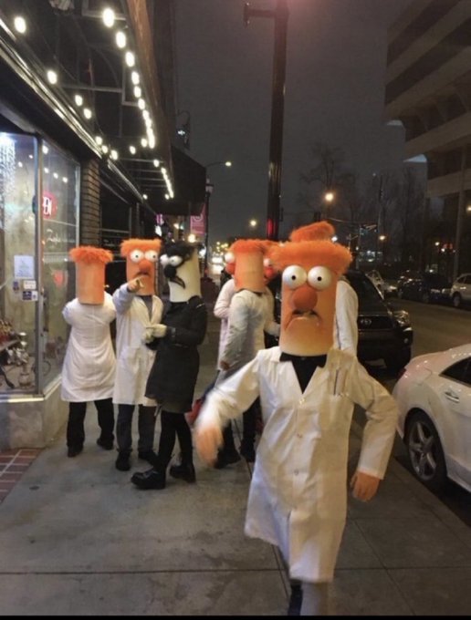 A nighttime street scene showing a group of people wearing orange Muppet-style mascot heads and white lab coats, with one person wearing a black suit standing among them on a city sidewalk.