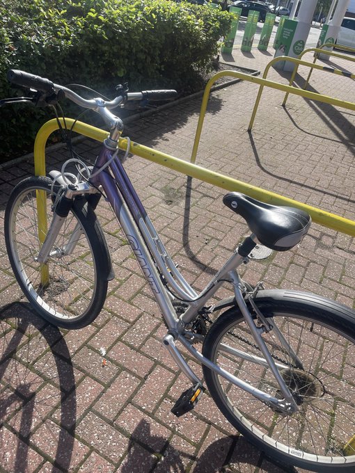 A silver and purple bicycle locked to a yellow metal bike rack on a brick pavement, with greenery and parking signs visible in the background.