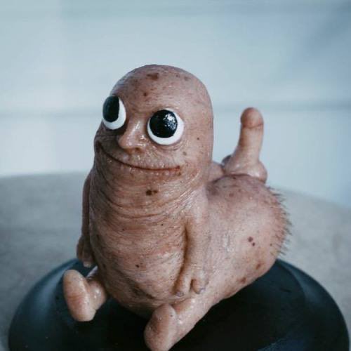 A sculpted, 3D-rendered figure resembling the "Dickbutt" internet meme—complete with googly eyes, stubble, and a curled tail—sits displayed on a black stand. The whimsical art piece ties to the post's suggestion that its creators could leverage the meme's viral fame to raise substantial funds for colon and prostate cancer charities.