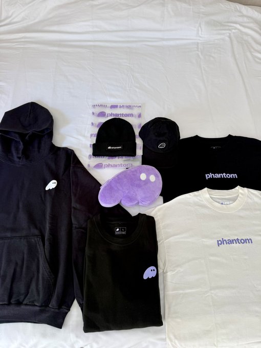 A collection of 'phantom' branded merchandise is laid out on a white sheet, including a black hoodie, white t-shirt, black tank top, beanie, and a purple plush item.