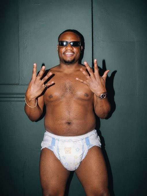 A shirtless man wearing sunglasses, a gold watch, and a gold bracelet, posing in a diaper decorated with teddy bears and stars against a dark green wall.