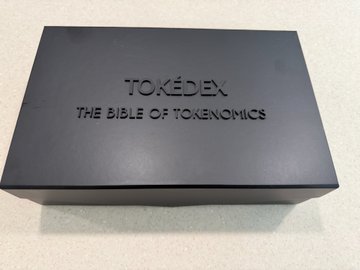 A sleek black box branded "TOKÉDEX - THE BIBLE OF TOKENOMICS" rests on a light surface, suggesting a premium product tied to the crypto industry's networking and advocacy efforts. The packaging likely represents a tokenomics resource or swag item connected to Robby and the broader crypto community referenced in the post.