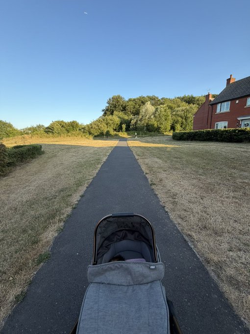 A person pushes a stroller down a paved path through a sunny, open grassy area beside a house. The image suggests a parent spending time with their child instead of chasing cryptocurrency airdrops.