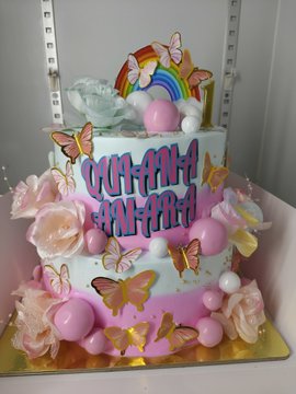 A two-tier pink and white birthday cake decorated with butterflies, roses, and a rainbow topper, with the text 'YUI-AMI 7TH BIRTHDAY' written on it.