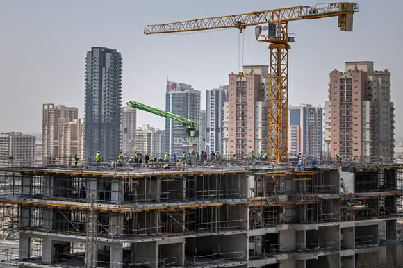 A construction site with workers and a yellow crane against a backdrop of high-rise city buildings, with a green boom lift visible on the upper floors.