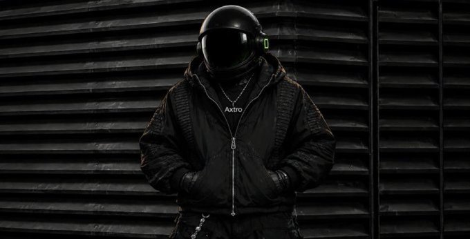 A person dressed in a black tactical jacket and a black motorcycle helmet stands against a dark, ridged wall, with 'Astro' visible on their shirt.