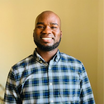 The image features a man wearing a blue plaid shirt, smiling at the camera with his hands placed behind his back. The background of the photo appears to be yellow, which contrasts nicely with the man's blue shirt. There are no texts or other objects in the image that seem to have any connection to the post about AI vulnerabilities and security reviews.