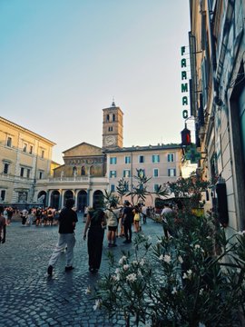 A bustling European piazza with historic buildings and a bell tower at golden hour, with a "CAFFAREL" sign visible on a building to the right, capturing the lively, timeless atmosphere that makes Rome such a vibe.