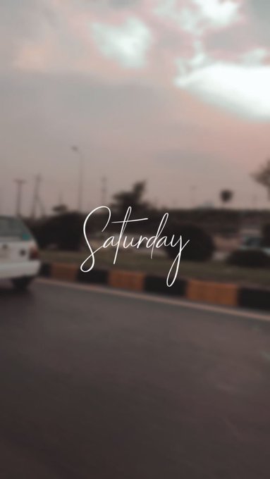 A blurred photograph of a street scene at dusk with the word 'Saturday' written in cursive white text overlaid in the center.