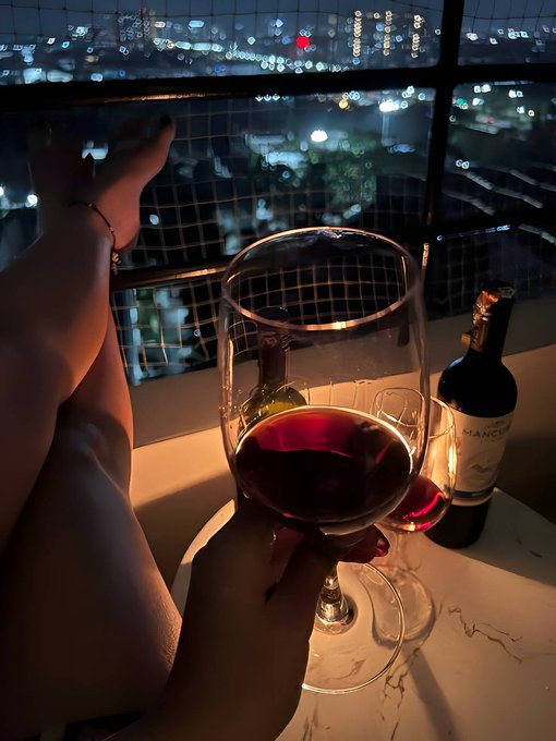A hand holds a glass of red wine on a marble ledge, with a wine bottle and city lights visible through a window at night.