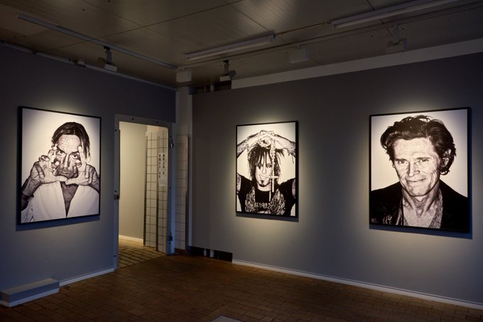 The image shows a gallery exhibition featuring three large black-and-white portrait photographs displayed on gray walls, illuminated by overhead spotlights in a minimalist white-walled space. The accompanying post references this as "iCONS of NFT 52icons," a genesis NFT collection from March 2022, showcasing portrait photography by Warren Finger (warrenhimself), suggesting these gallery prints represent physical or digital art tied to the NFT series.