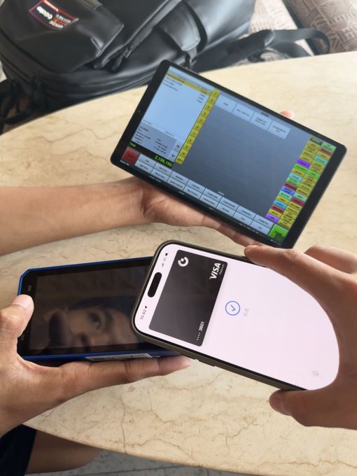 A person uses a tablet displaying a point-of-sale system to process a contactless payment via a Visa card held over a smartphone, capturing the convenience of the GateCard digital payment experience. The scene illustrates the final transaction on a trip, tying into the post's "Last Day on the Island" theme as customers wrap up their visit with a quick, seamless payment.