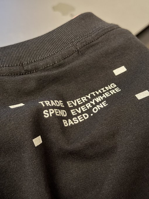 Close-up of a dark gray garment featuring the white printed text 'TRADE EVERYTHING SPEND EVERYWHERE BASED .ONE'.