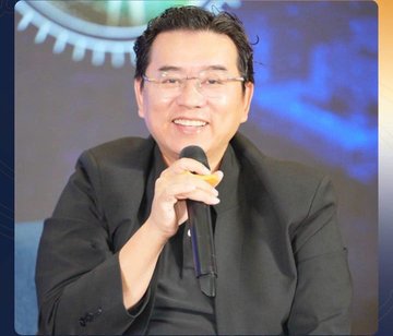 A photograph of a smiling man wearing glasses and a dark suit, holding a microphone while speaking on stage.
