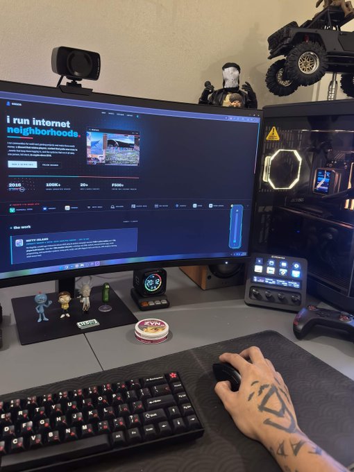 A tattooed hand operates a mouse on a desk setup featuring a large monitor displaying the 'i run internet neighborhoods.' website, alongside a mechanical keyboard, gaming PC, and small figurines.