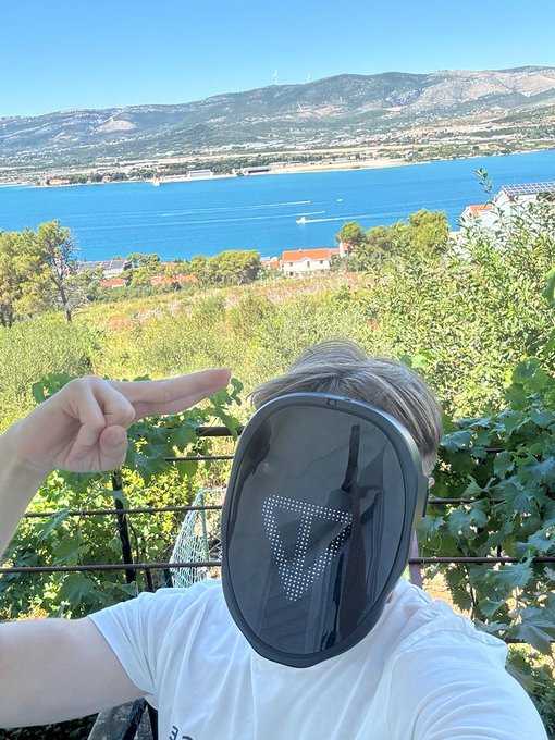 A person wearing a visor and white t-shirt poses with their hand near their head against a scenic coastal backdrop featuring blue water, boats, and mountains.