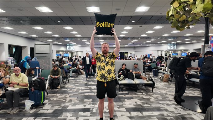 A man stands in a busy airport terminal holding up a black pillow with yellow 'Solflair' text above his head.