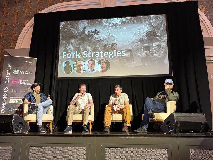 Four men sit on a stage holding microphones during a panel discussion titled "Fork Strategies," with an "Attendees" banner showing sponsors like NYDIG and Human Rights Foundation for the Consensus Edition.