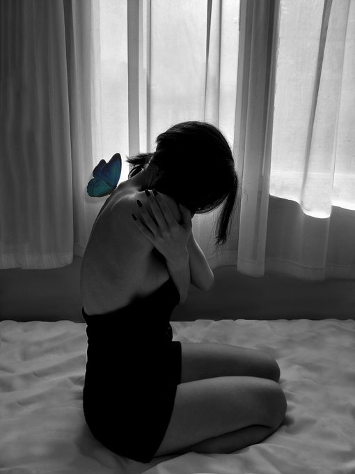 A black-and-white photograph of a woman with dark hair in a black dress sitting on a bed with her hand near her face, accented by a small blue butterfly near her shoulder.