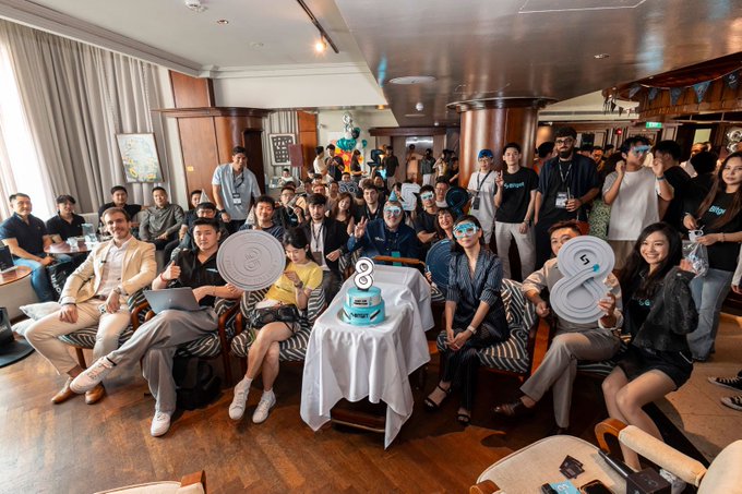 A large group of people gathered indoors at what appears to be a corporate or community event, with attendees posing with large circular signs and a birthday cake.