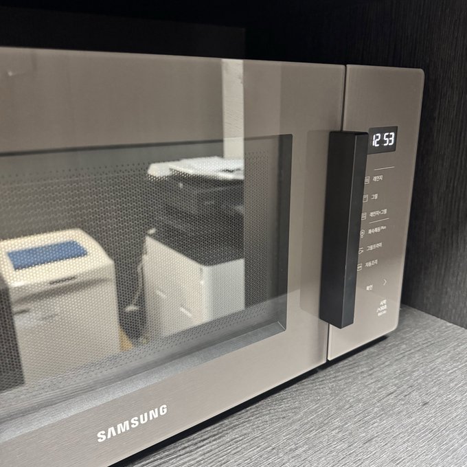 A Samsung microwave with a digital display showing "12:53" is shown in an office setting, with reflections of office furniture visible on its glass door.