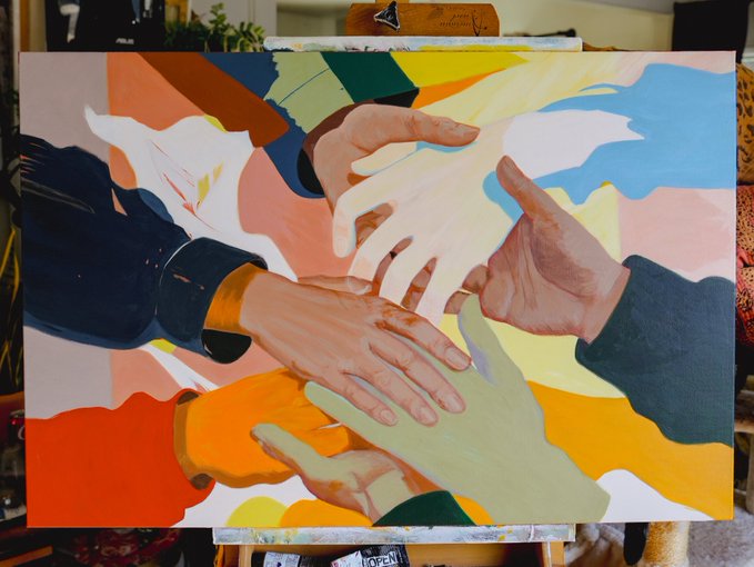 A vibrant painting on an easel depicts multiple hands of various skin tones reaching toward and touching each other against a colorful abstract background.