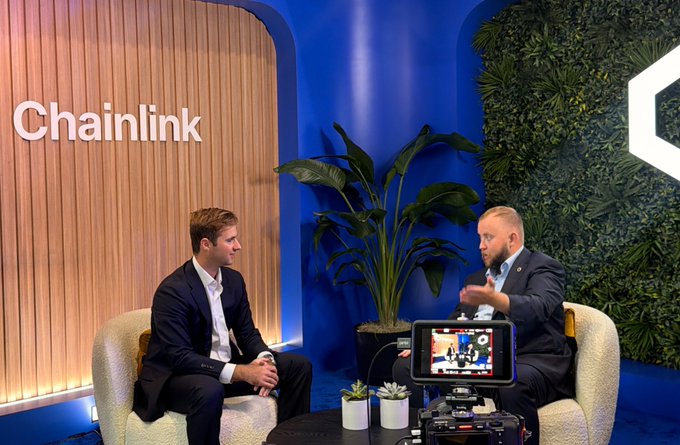 An interview setting with two men seated in armchairs in front of a 'Chainlink' sign and a blue plant wall, filmed by a camera in the foreground.